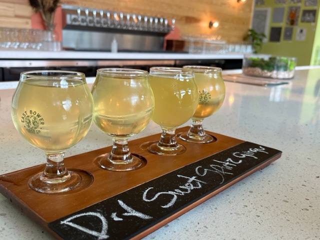 four small flight glasses of cider on a counter; taps in backgrousn