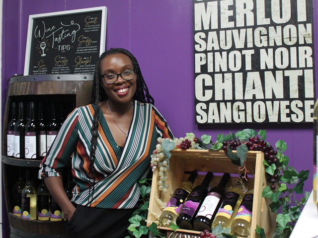 woman standing beside a case of wine, sign on the wall, wine bottles behind her
