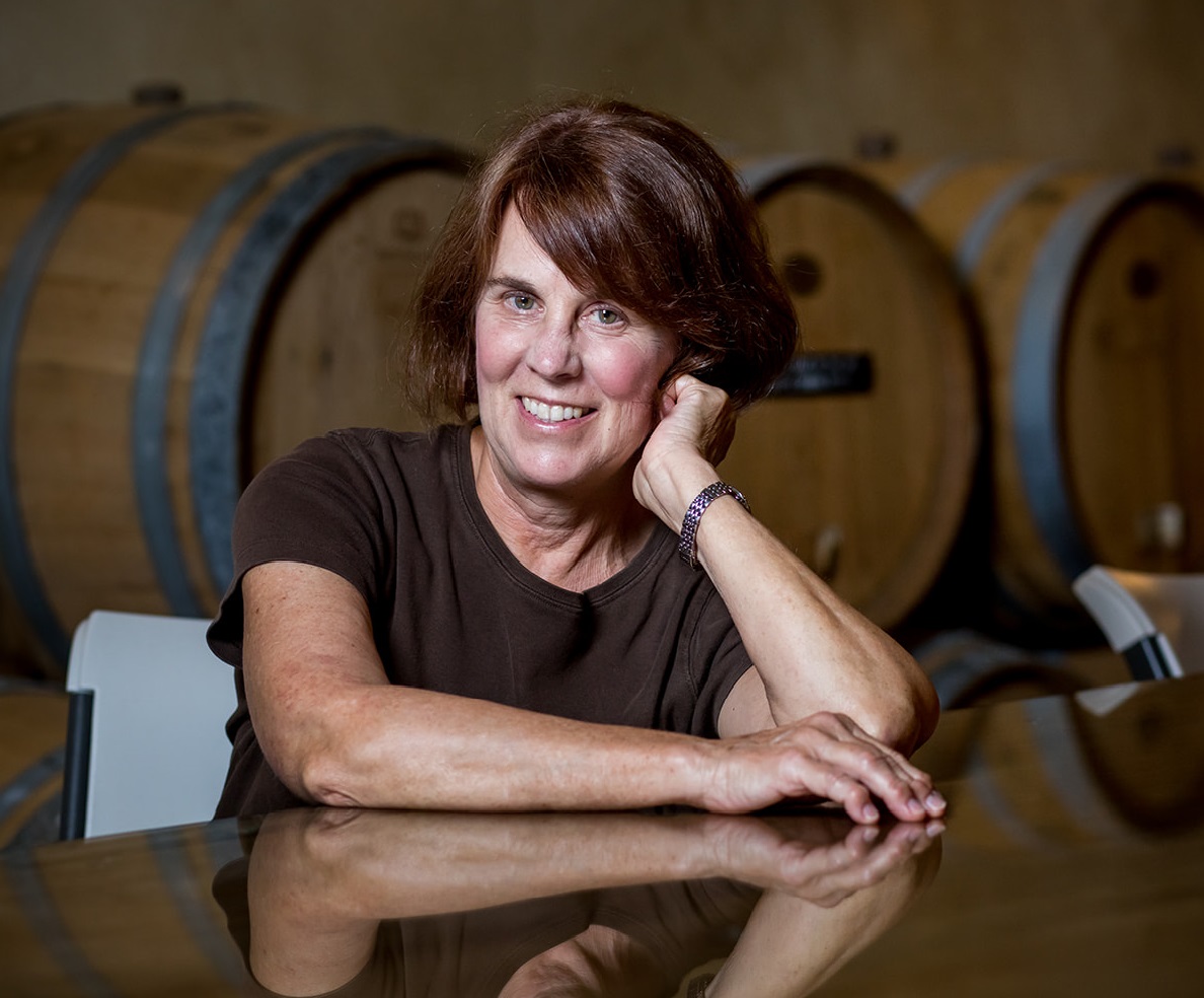 Natalie, seated at table with one arm out and hand supporting her head. wine barrels in the background