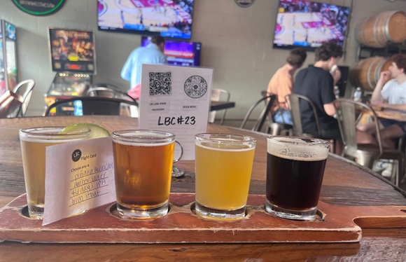 a flight of beers on a table, with tv monitors in the background