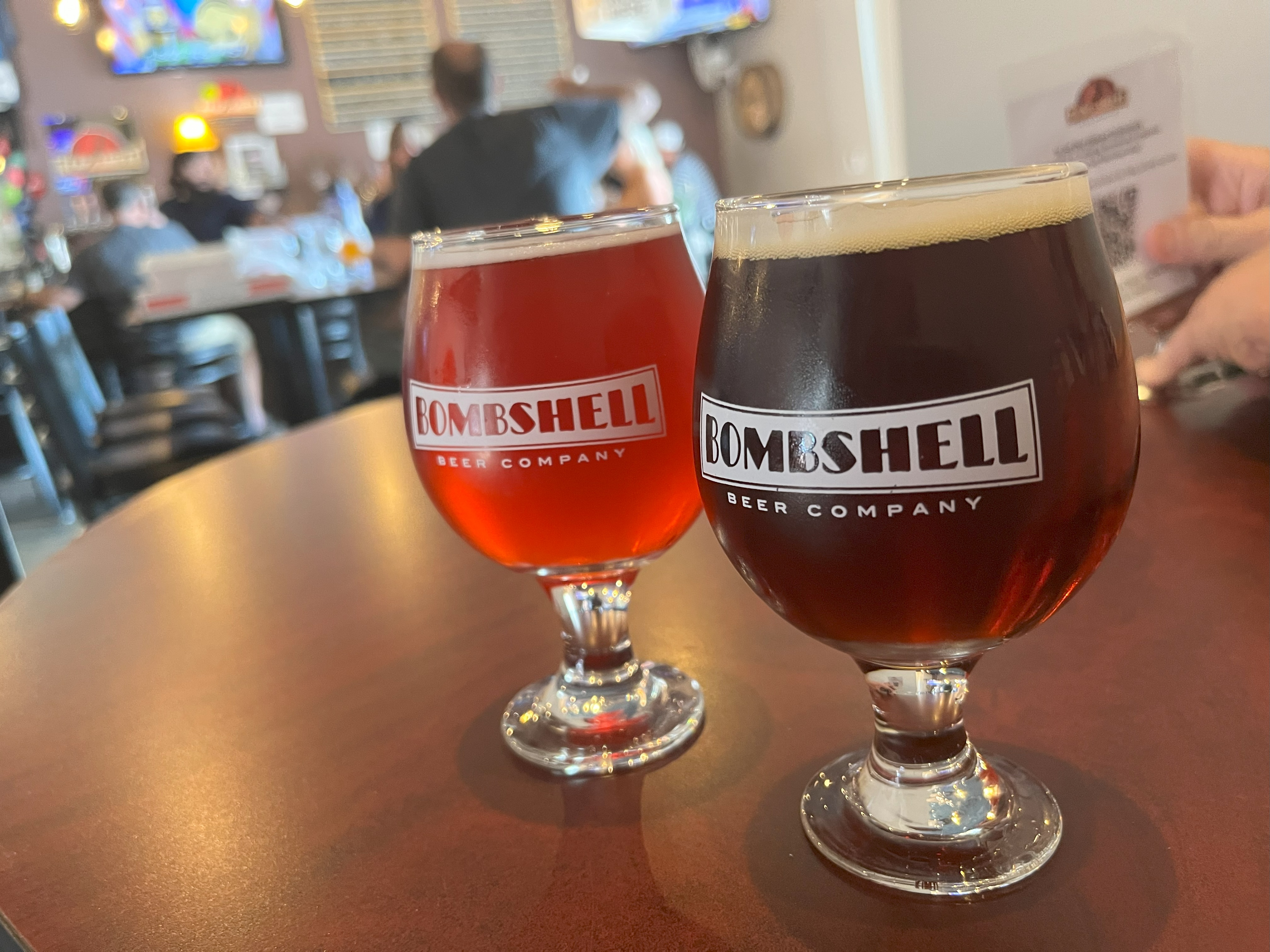 Two Bombshell Beer glasses with beer, on table, with taproom in background