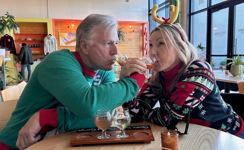 man and woman sipping ciders with arms crossed, and wearing christmas sweaters