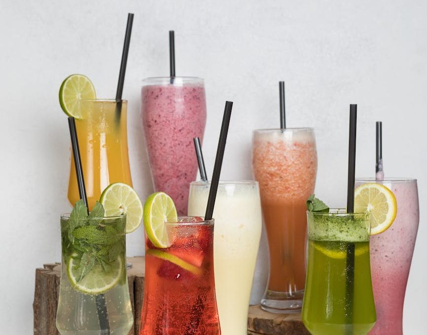 eight glasses with various colors of drinks and straws